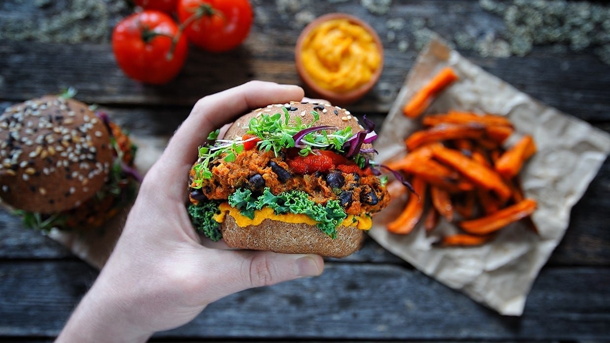 Persona sosteniendo una hamburguesa vegana con vegetales de fondo.